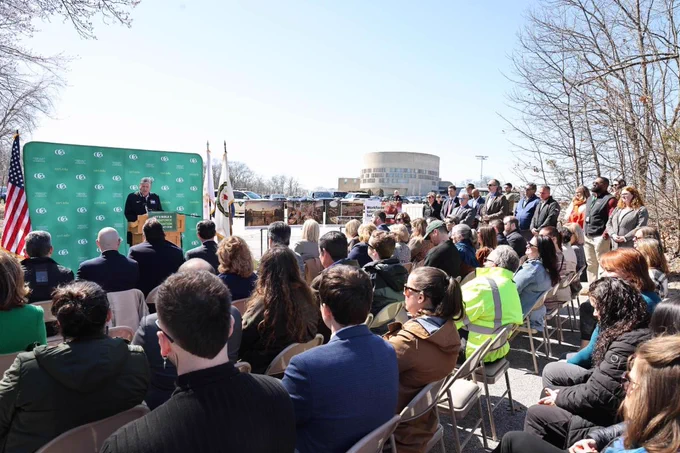 Outdoor event with Governor Dan Mckee addressing a seated audience against a green backdrop and American flags on a clear day.