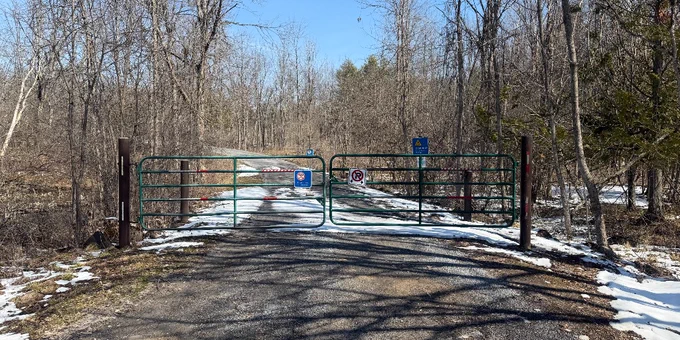a road with a green gate closed at the end of it, with snow appearing in large spots on the road.