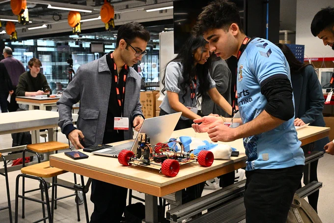 Students collaborating on a robotics project with a laptop and a small vehicle with red wheels on a workshop table.