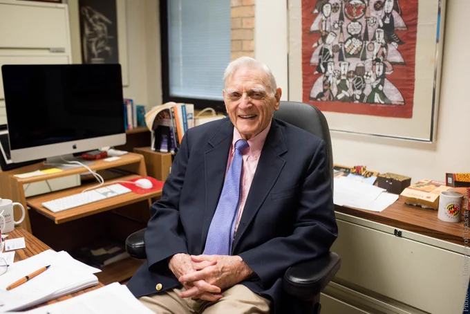 John B. Goodenough sitting in an office chair at a desk, smiling at the camera, with a computer and books on the desk. 