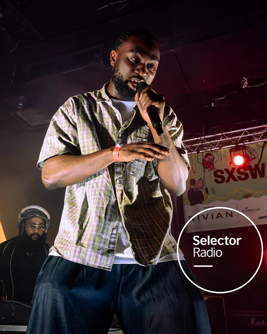 Kofi Stone holding a microphone mid-speech on a dimly lit stage at SXSW, with a DJ in the background. Selector Radio is written in white in a transparent circle in the bottom left of the image.