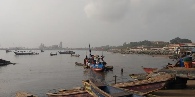 Fishers guide a canoe across a harbor. The sky is overcast, and buildings appear on the horizon. Image credit: Adelina Mensah