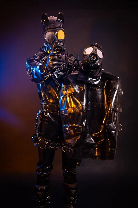 GimpSimp in a heavy rubber suit, heavy harness and hooded gas mask. He is pointing at the viewer holding another gas mask and a rubber body binder