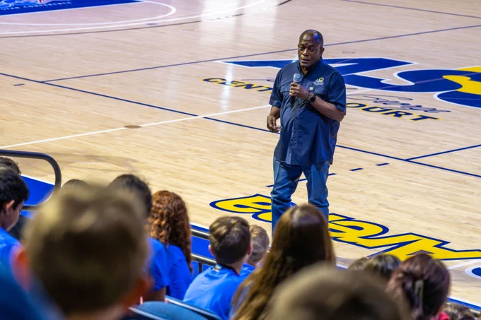 A man stands on a basketball court, speaking into a microphone. In the foreground, a group of seated people wearing blue shirts listens attentively.