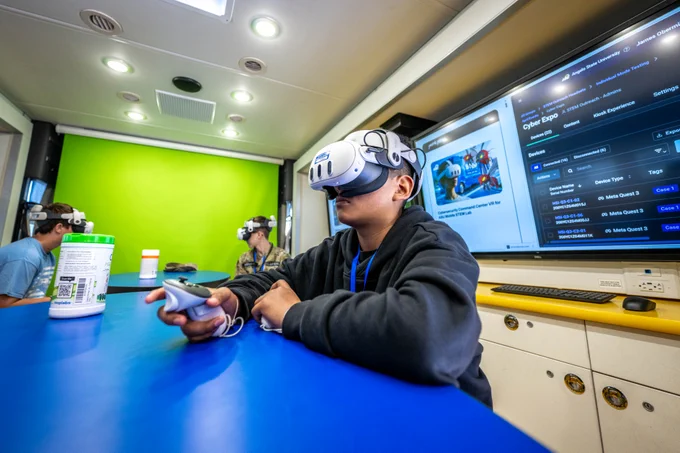 Three people wearing virtual reality headsets sit at a blue table in a room with a green screen background. One individual is in the foreground holding a controller, while two others are visible in the background. A large monitor displaying text and graphics is mounted on the wall to the right.
