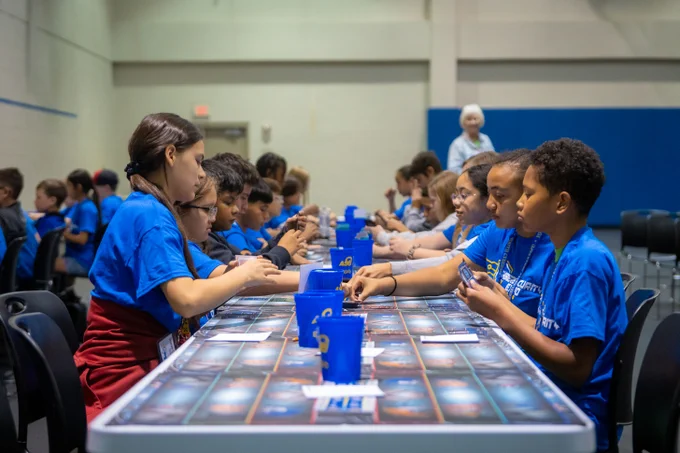 Children in blue shirts sit around a long table playing a card game, each focusing on their hands. The tabletop is covered with a gaming mat and has blue cups scattered along it. 