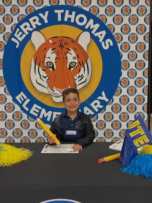Young boy sitting at a table holding a large pencil in front of a sign for Jerry Thomas Elementary.