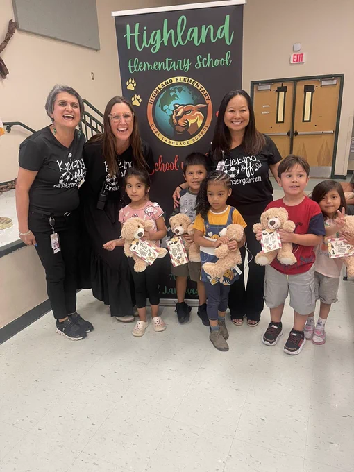 Group of 5 students holding teddy bears with three educators in front of a sign for Highland Elementary School.