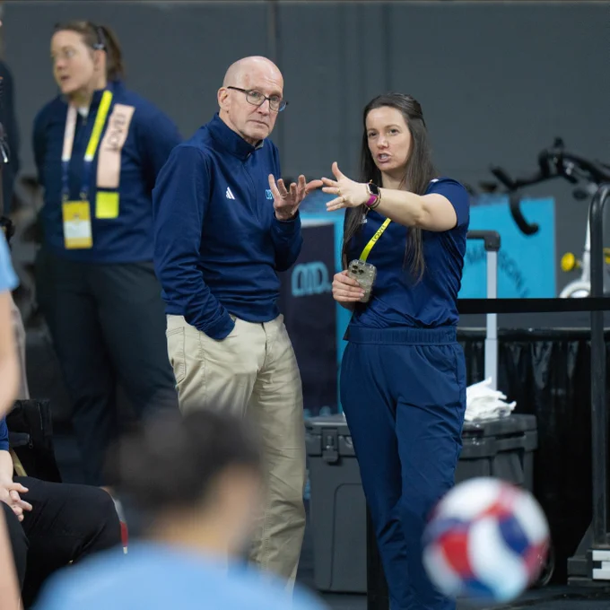 A person with an official credential gestures while talking to another person in a sports setting. A volleyball is visible in the foreground. Other individuals and equipment are present in the background.