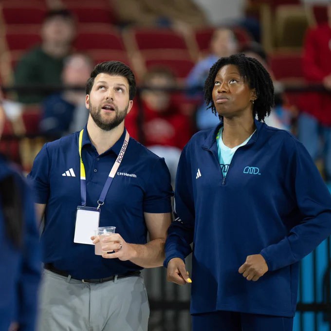 Two people are standing together in a gymnasium with a blurred audience in the background. One person is wearing a navy shirt with an "LOVB" lanyard and holding a drink, while the other is in a blue players jacket. Both are looking up attentively.