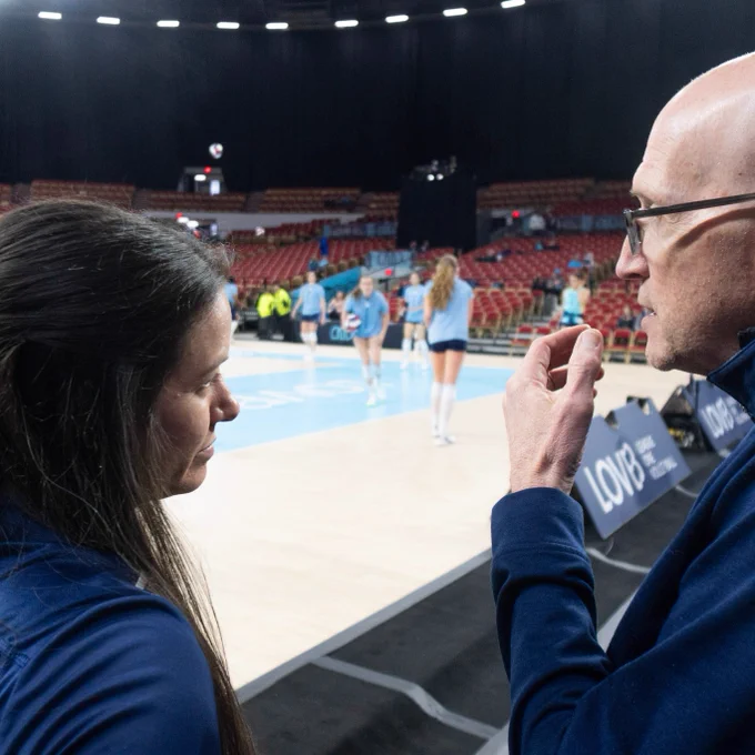 Two people in blue team jackets discussing on the sidelines of a volleyball court. 