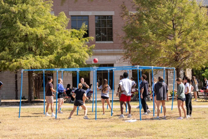 Group of people playing four square volleyball on a blue metal frame outdoors near a brick building.