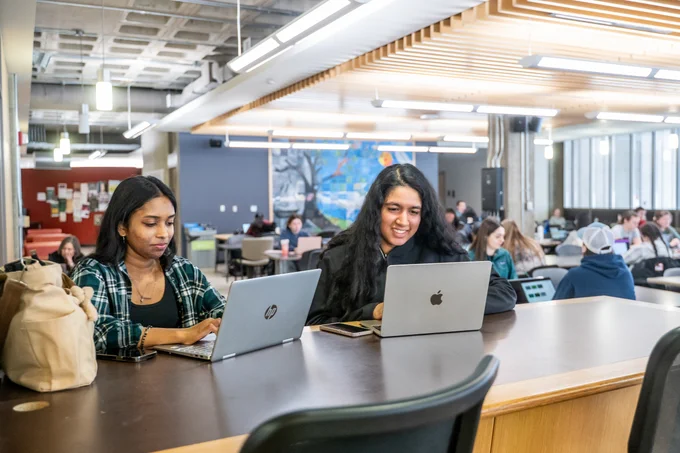 Two people sitting at a table using HP and Apple laptops in a busy, open-plan study area. A colorful mural is visible on the wall in the background, with several other people seated and studying.
