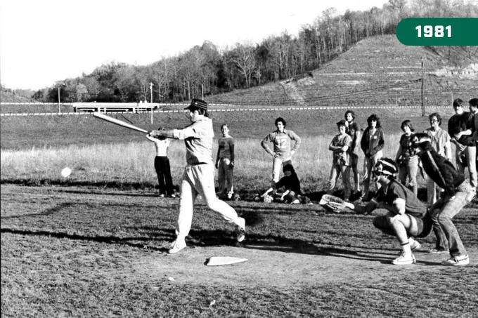 A black and white archival photo from 1981 showing a Heritage College student mid-swing during a baseball game on a grassy field, with classmates watching from the sidelines and hills in the background.