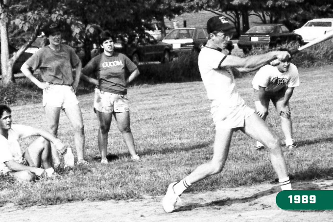 A black and white archival photo from 1989 featuring a student in a baseball cap and glasses swinging a bat at home plate while other students watch from the grass.