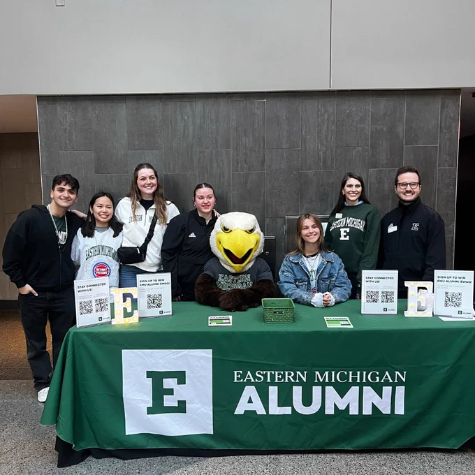 A group of people, including someone in an eagle mascot costume, stand behind a table with an "Eastern Michigan Alumni" tablecloth. The table displays flyers and QR codes. The background features a neutral-colored wall.