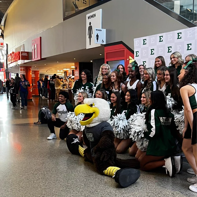 A group of cheerleaders pose with a person in an Eastern Michigan University mascot costume at an indoor event. There's a mall-style directory sign above them and a store called AT&T visible in the background.