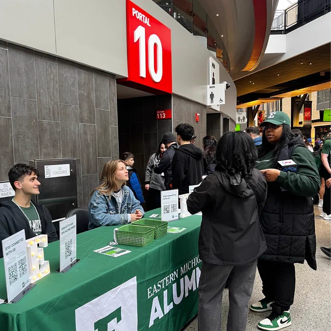 A group of individuals are gathered at a table covered with a green "Eastern Michigan Alumni" cloth inside a venue near Portal 10. There are informational signs on the table, and people appear to be conversing. Attendees are wearing casual attire, and the scene seems lively and engaging.