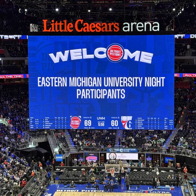 The image shows a basketball game at Little Caesars Arena with a large scoreboard displaying "Welcome Eastern Michigan University Night Participants." It shows the Detroit Pistons leading the Philadelphia 76ers with a score of 69 to 60 and 5:56 minutes remaining in the fourth quarter. Spectators fill the stands around the court.