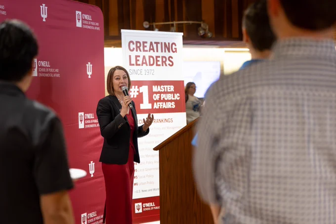 Dean Sian Mooney speaks into a microphone at a podium, addressing an audience. The backdrop features "O'Neill School of Public and Environmental Affairs" with an Indiana University logo. A sign highlights the program's ranking as "#1 Master of Public Affairs.