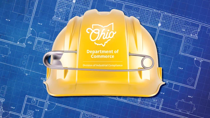 A yellow hard hat with the Ohio Department of Commerce logo, featuring an outline of Ohio and text "Division of Industrial Compliance," rests on blue architectural plans. A large silver safety pin is clipped onto the hard hat.