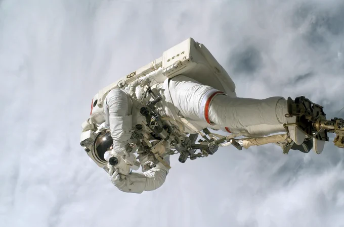 A photo of an astronaut tethered by their feet and holding a camera in front of their helmet as if taking a photo. The background features a bright, cloudy sky.