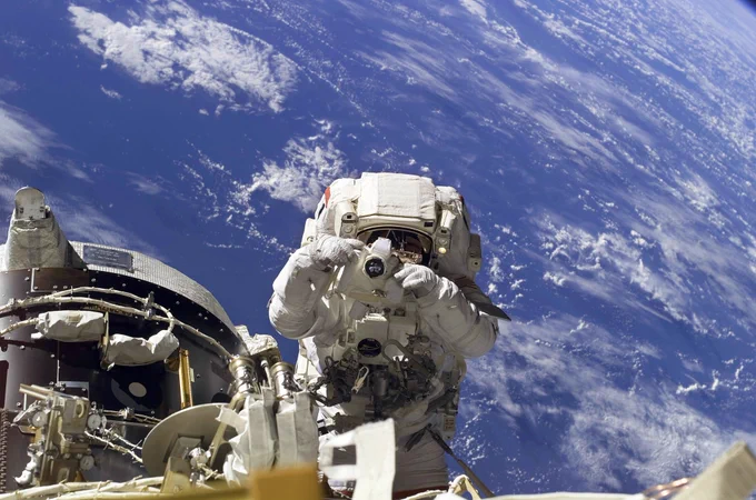 A photo of an astronaut in a white spacesuit using a camera to take a photo while floating in space. The earth is visible behind them.