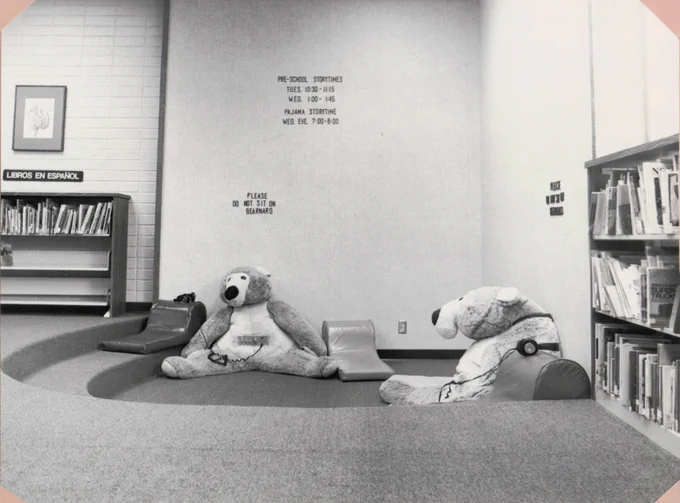 black and white photo of library interior. books on shelves on either side of photo. centered are two large bears on the recessed floor with headphones plugged in to audiocassette players built into the bears' middle. 