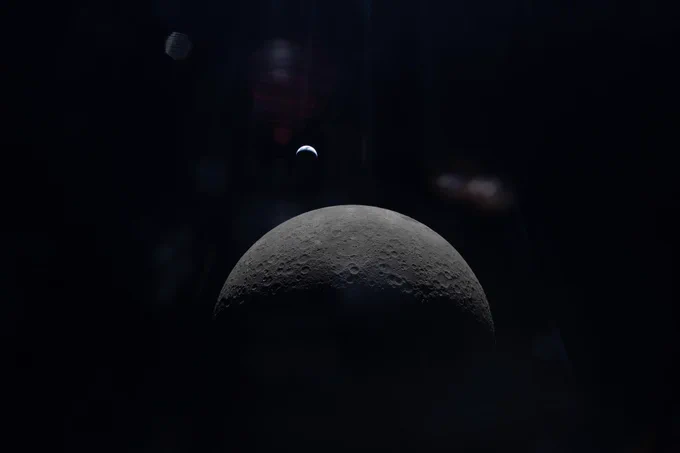 The top part of the Moon is illuminated. The gray cratered surface stands out against the blackness of space. The Earth appears in the far distance as an upside-down crescent moon shape. There is some lens flare in the top part and center of the image. Credit: NASA