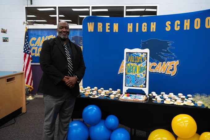 The new principal at Wren High School standing with the schools banner with a welcome sign and cupcakes behind him. 