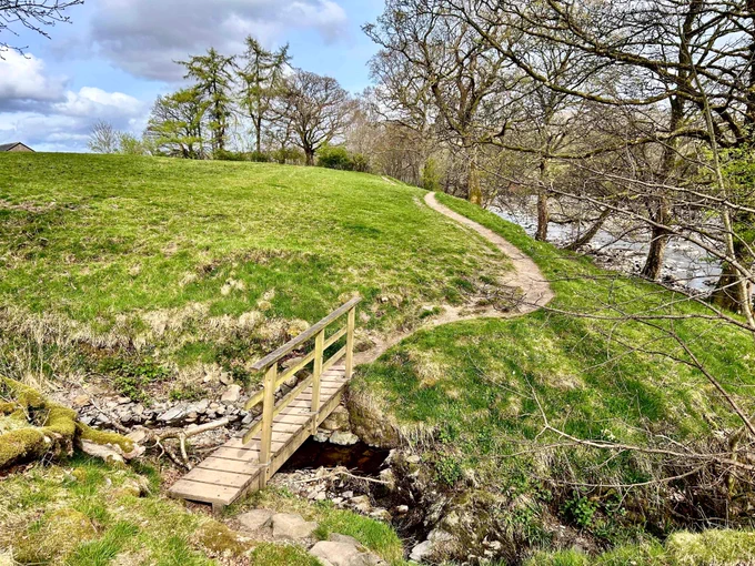 Narrow wooden footbridge crossing a small creek with a winding dirt path leading up a grassy hill under a partly cloudy sky.