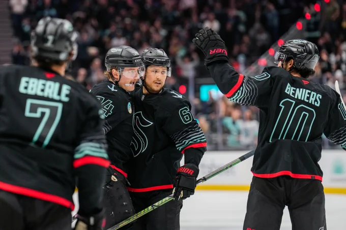 a photo of Seattle Kraken defender Brandon Montour with his arms wrapped around forward Bobby McMann. Matty Beniers and Jordan Eberle are skating in to hug the duo.