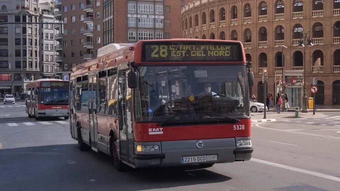 Autobús urbano rojo (línea 28) detenido en una avenida frente a un edificio de arcos, con otro autobús y peatones cerca.