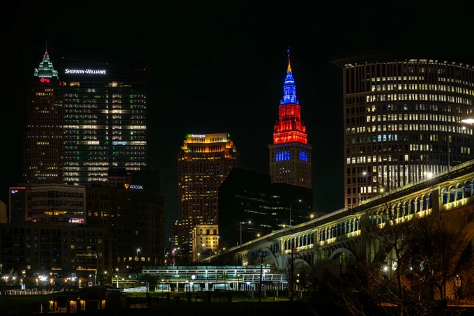 A nighttime view of the Cleveland city skyline from the Flats area. The top of Terminal Tower, the second building from the right, is illuminated blue and orange. A bridge stretches away from the skyscrapers to the right. The sky above the buildings is dark. Credit: NASA/Jef Janis