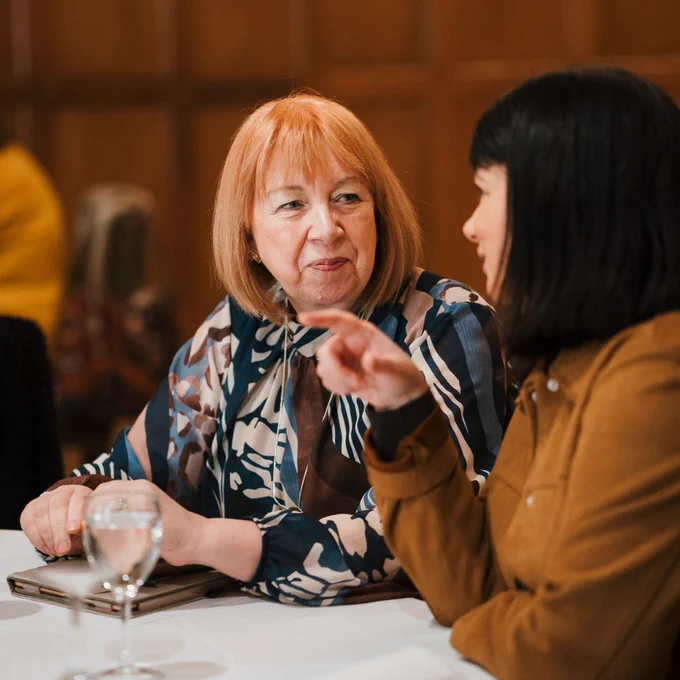Two women engaged in a focused conversation at a dining table with glasses of water in a warmly lit room.