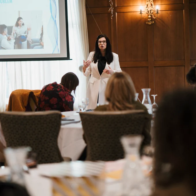 Woman in white blazer presenting to an attentive audience in a wood-paneled conference room with a projection screen.