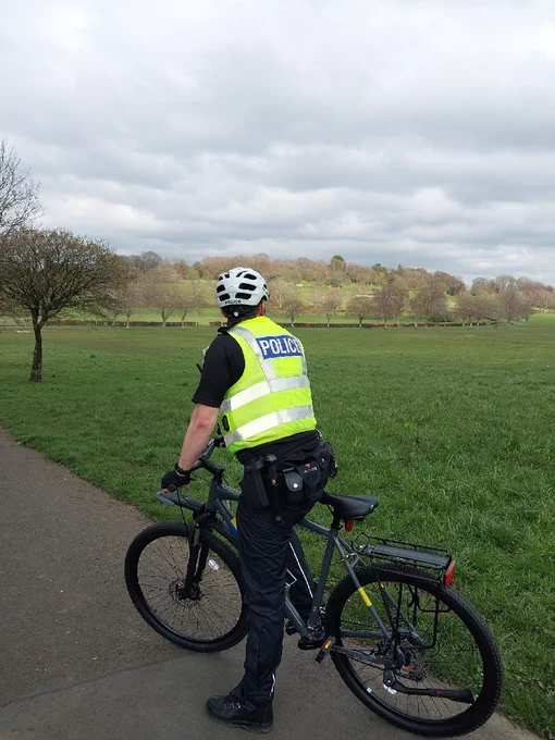 Police officer in high visibility uniform on cycle patrol in a large park area.