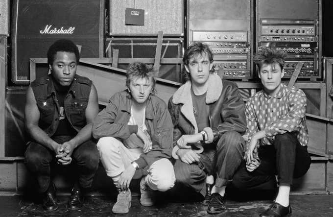 Black-and-white promotional photo of Big Country. From left to right: Tony Butler (bass), Bruce Watson (guitar), Mark Brzezicki (drums), and Stuart Adamson (lead vocals and guitar), seated together in front of stacked amplifiers.