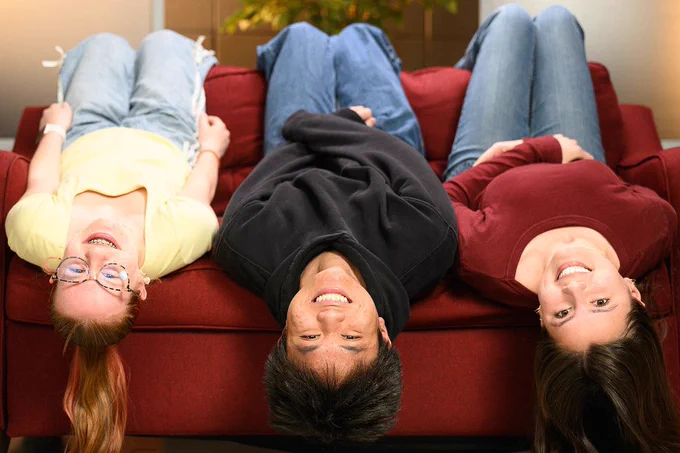 Three students hang upside down from the edge of a red couch.