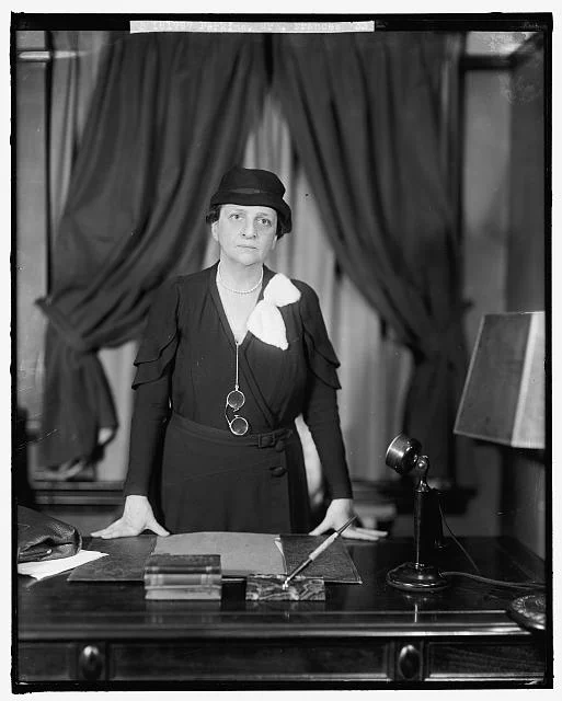 A photo of Frances Perkins standing behind a desk. Credit: Library of Congress.