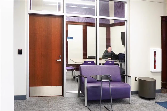 A person is seated at a desk inside a room with glass walls, visible from a hallway. The room has a sign labeled "Meeting Room." In the foreground, there are purple chairs and a small round table.
