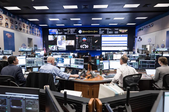 View inside of NASA's Mission Control Center. In the foreground, four people sit at desks with their backs to the camera. They each have multiple screens in front of them. There are multiple rows of desks in front of them and on the far wall are large screens with even more data. Credit: NASA