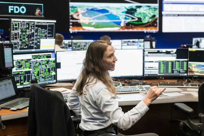 A woman sits at a desk at Mission Control. She is angled to the right with her arm outstretched as she talks to someone. On the desk in front of her are multiple screens with data and visualizations. In the background, on the far wall, are even more screens. Credit: NASA