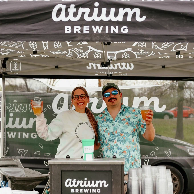 Two people stand behind an Atrium Brewing booth holding plastic cups of beer under a branded canopy tent.