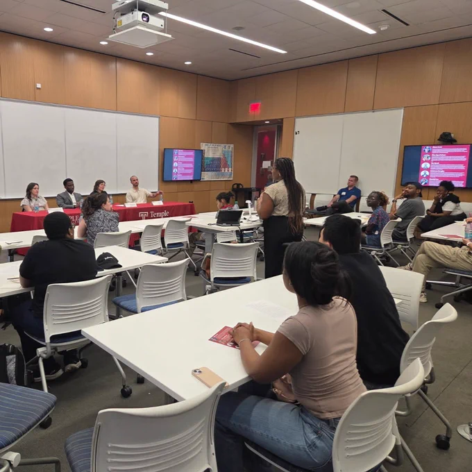 A classroom setting with students seated at tables facing a panel of individuals. One individual standing in the center of the room is moderating the panel. Attendees are listening attentively.