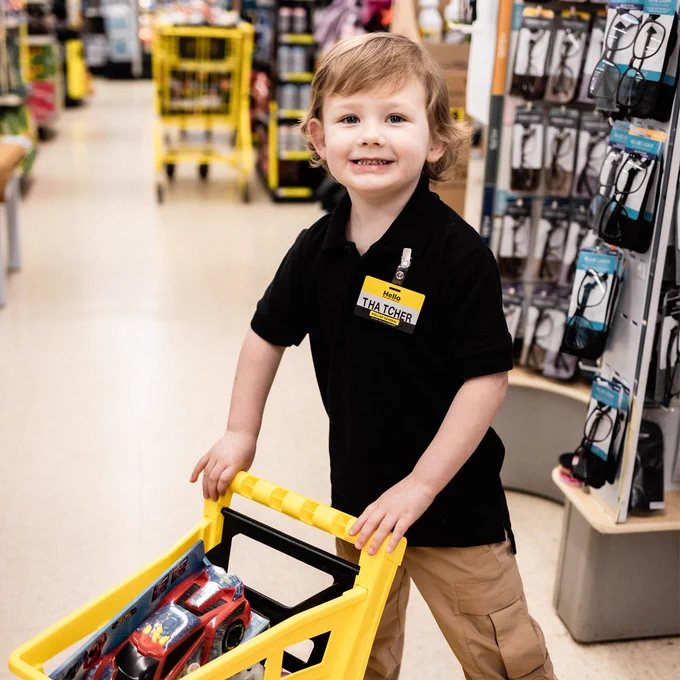A young boy with a \"Hello, THATCHER\" name tag smiles, pushing a yellow shopping cart with a toy car inside. He's in a store aisle with products on shelves, dressed in a black polo shirt and khakis.