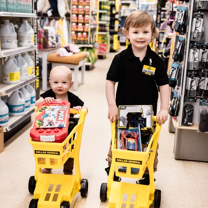 Two young children in a Dollar General. The younger child is sitting in a small, yellow shopping cart filled with a red \"MAX 253 PIECES@1\" toy. An older child in a black shirt, wearing a name tag that says \"Hello THATCHER@2\", is also holding a yellow cart.