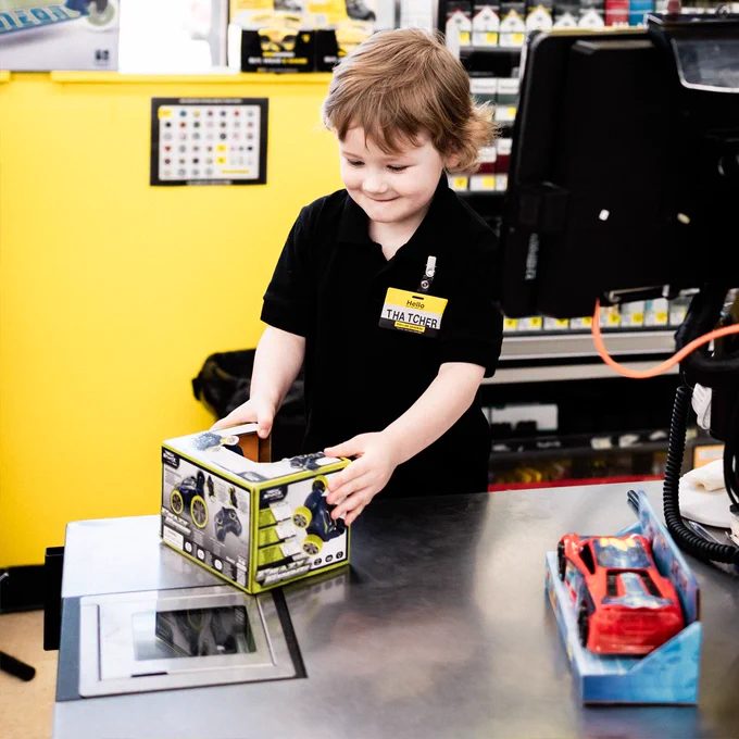 A young child, \"Hello THATCHER\", in a black polo shirt, examines a toy box at a store checkout counter. A toy car sits nearby. The background is a bright yellow wall with displayed items.