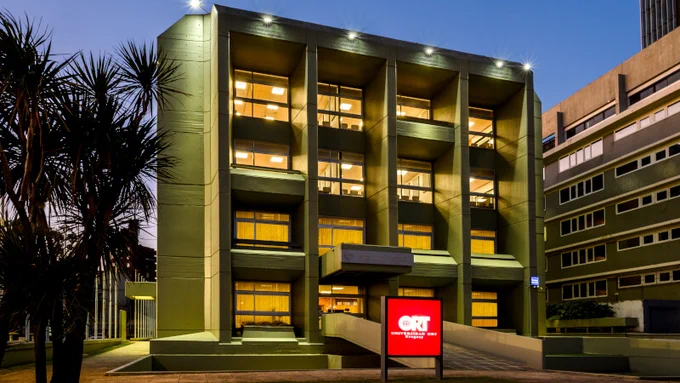 Fachada iluminada al atardecer de edificio universitario con ventanas encendidas y cartel rojo en primer plano