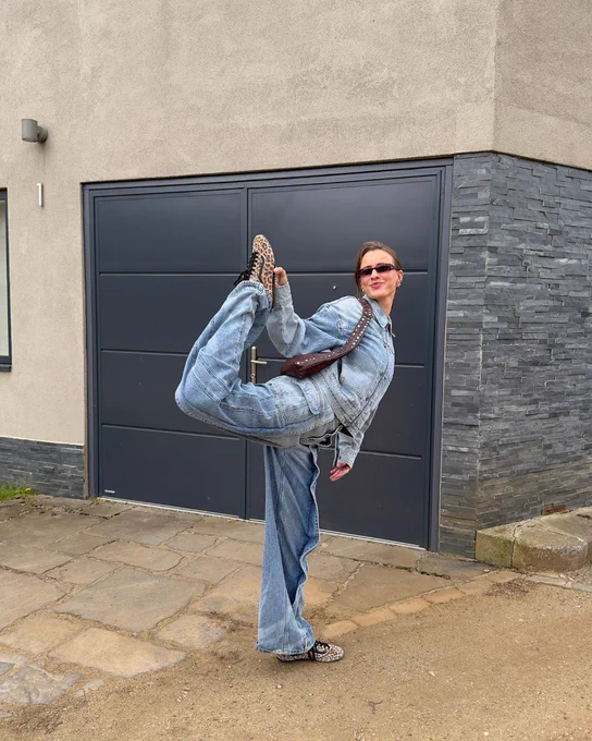 An image of a person wearing a pair of leopard print Vans Super Lowpro trainers with a denim outfit whilst standing on one leg in front of a stone building.